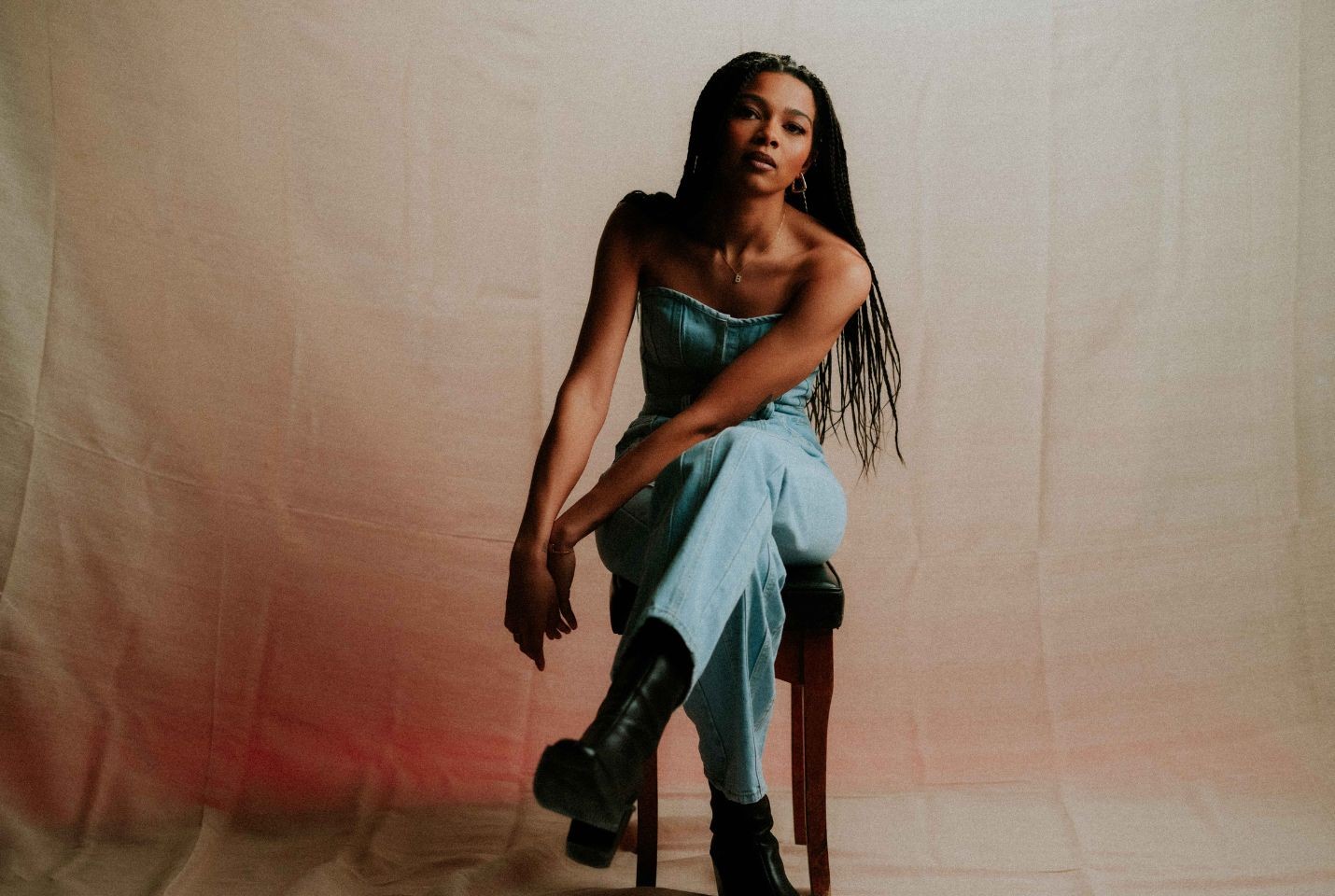 A woman with ling braided hair in a denim jumpsuit sat on a stool